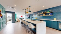 Located atop 2061 Rue Stanley in the heart of Montréal, this rooftop lounge and terrace is a boldly designed communal space that seamlessly blends vibrant interiors with open-air living. The interior lounge features light herringbone hardwood flooring, floor-to-ceiling windows, and teal-accented architectural columns that flood the space with natural light and sweeping city views. A plush green velvet sofa, curved accent chairs in warm tones, and curated artwork contribute to the eclectic yet sophisticated atmosphere. The fully equipped kitchen is a standout feature, showcasing a lengthy marble-topped island with boucle bar seating, deep teal cabinetry, a vivid blue mosaic tile backsplash, open wood shelving, and playful pendant lighting. A separate dining area offers a striking contrast with its painted blue brick accent wall, chevron parquet flooring, and bold red pendant light above a sleek table. The expansive outdoor terrace wraps around the building and is divided into distinct zones, including lounge areas centered around fire pit tables with plush seating, high-top dining tables with colorful chairs, and a BBQ area with wicker patio sets — all set against a backdrop of Montréal's iconic downtown skyline and Mount Royal.