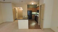 This single-floor residence showcases an open-concept kitchen and living area designed for functional everyday living. The kitchen is outfitted with warm honey-toned wood cabinetry that extends to the upper walls, providing generous storage throughout. Dark countertops top both the perimeter cabinetry and a central peninsula, which features a double-basin sink and serves as a natural divider between the kitchen and the adjoining carpeted living space. Pendant lights hang above the peninsula, adding focused task lighting with a touch of style. Black appliances, including a full-size refrigerator, range, and microwave, complement the darker countertop tones. The kitchen floor is finished in hardwood-style flooring, contrasting with the plush carpet that extends into the living area. Sage green accent walls in the kitchen add a subtle pop of color against the neutral cream tones throughout the rest of the space. A hallway visible from the main area leads to additional rooms, with multiple doors suggesting well-separated living quarters. The layout feels cohesive and efficiently organized, making practical use of the available footprint.