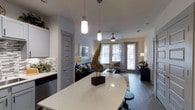 This 1-bedroom, 1-bathroom residence is thoughtfully designed with an open-concept floor plan that maximizes the use of space and promotes a seamless flow between living areas. The kitchen is a standout feature, showcasing white shaker-style cabinetry, a decorative mosaic tile backsplash, stainless steel appliances, and a generously sized curved quartz countertop island that doubles as a breakfast bar with seating. Pendant lighting with industrial-style fixtures hangs above the island, adding both function and visual interest. The kitchen opens directly into the living area, which is bathed in natural light from large windows and a set of glass doors that invite outdoor views and fresh air into the space. A ceiling fan overhead enhances comfort throughout the room. Rich wood-tone flooring runs throughout, providing warmth and continuity across the open layout. Soft, neutral wall tones complement the modern aesthetic, while patterned curtains and curated décor accents add personality to the space. The overall ambiance is modern, welcoming, and move-in ready, with quality finishes and thoughtful design details evident throughout.
