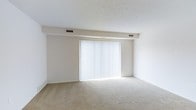 This 1-bedroom, 1-bathroom residence at The Garden is a single-floor home designed for comfortable, everyday living. The layout is thoughtfully arranged to maximize functionality while maintaining a clean, neutral aesthetic throughout. The living area is finished with plush wall-to-wall carpeting in a warm beige tone, creating a cozy and inviting atmosphere. A large sliding glass door framed by vertical blinds serves as the focal point of the main living space, allowing abundant natural light to fill the room while providing direct access to the outdoors. The white walls and crisp ceiling with recessed HVAC vents contribute to a bright, airy feel. The architectural details are simple and understated, with a subtle ceiling beam accent that adds a modest structural character to the space. Overall, the residence presents a clean canvas that is easy to personalize, making it a practical and welcoming home. Its straightforward design, natural light, and neutral palette combine to create a livable space well-suited for a range of lifestyles.