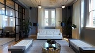 This stylish single-floor space exudes a refined, contemporary elegance rooted in thoughtful design. The main living area serves as the centerpiece, showcasing dark charcoal walls that create a dramatic yet inviting atmosphere, beautifully contrasted by warm hardwood flooring and an abundance of natural light streaming through tall windows dressed in flowing cream drapery. A tufted white sofa anchors the seating arrangement, complemented by a striking mirrored and gold circular coffee table that adds a glamorous focal point to the room. Coordinating white tufted ottomans flank the space, providing both style and function. Overhead, a sculptural chandelier with organic, amber-toned elements draws the eye upward, accompanied by sleek pendant lighting that enhances the overall illumination. Black floor lamps with bold shades add layers of warm ambient light throughout. Lush tropical plants positioned throughout the room introduce a vibrant natural element that softens the space's sophisticated palette. The interplay of deep wall tones, metallic accents, crisp white upholstery, and organic greenery gives this space a polished, editorial quality ideal for both relaxed gatherings and upscale entertaining.