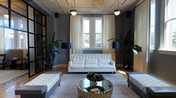 This stylish single-floor space exudes a refined, contemporary elegance rooted in thoughtful design. The main living area serves as the centerpiece, showcasing dark charcoal walls that create a dramatic yet inviting atmosphere, beautifully contrasted by warm hardwood flooring and an abundance of natural light streaming through tall windows dressed in flowing cream drapery. A tufted white sofa anchors the seating arrangement, complemented by a striking mirrored and gold circular coffee table that adds a glamorous focal point to the room. Coordinating white tufted ottomans flank the space, providing both style and function. Overhead, a sculptural chandelier with organic, amber-toned elements draws the eye upward, accompanied by sleek pendant lighting that enhances the overall illumination. Black floor lamps with bold shades add layers of warm ambient light throughout. Lush tropical plants positioned throughout the room introduce a vibrant natural element that softens the space's sophisticated palette. The interplay of deep wall tones, metallic accents, crisp white upholstery, and organic greenery gives this space a polished, editorial quality ideal for both relaxed gatherings and upscale entertaining.