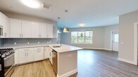 This well-appointed two-bedroom, two-bathroom residence at 5 Lakeshore Center in Bridgewater, MA offers a thoughtfully designed single-floor layout with modern finishes throughout. The open-concept kitchen and living area serves as the heart of the home, featuring white shaker cabinetry, a subway tile backsplash, stainless steel appliances, and a large center island with a light stone countertop and pendant lighting above. Hardwood-style flooring flows through the main living area, which is filled with natural light from a generous window offering outdoor views of the surrounding community. The spacious primary bedroom is carpeted for comfort and includes a private ensuite bathroom fitted with wood-tone tile flooring, a glass-enclosed walk-in shower, and white cabinetry. The second bedroom is equally well-sized, also carpeted, and features a window with outdoor views. The second bathroom mirrors the refined aesthetic of the primary, with tile flooring, a soaking tub with tiled surround, a white vanity, and a framed mirror. A clean, neutral color palette of soft grays and whites ties the space together, lending a bright and contemporary ambiance throughout.