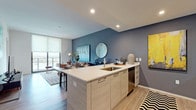 This stylish, single-floor residence is designed with a modern aesthetic and an open-concept layout that seamlessly connects the kitchen, dining, and living areas. The kitchen serves as a central hub, featuring a peninsula with a white quartz countertop, an undermount sink, light wood cabinetry, and a full suite of stainless steel appliances including a refrigerator, electric range, over-range microwave, and dishwasher. The open living and dining area is anchored by a bold slate-blue accent wall and light wood-plank flooring, creating a sophisticated, contemporary atmosphere. Floor-to-ceiling windows and sliding glass doors flood the space with natural light and frame sweeping views of the surrounding cityscape and waterways. The bedroom continues this elevated aesthetic with plush carpeting, a dedicated balcony with outdoor seating, and generous roller shade window treatments. The bathroom is finished with large-format white tile flooring, light wood cabinetry paired with a white countertop vanity, a glass-enclosed walk-in shower, and a dark accent wall that adds depth to the clean, minimal design. Recessed lighting throughout the residence enhances the bright, airy ambiance of this well-appointed urban home.