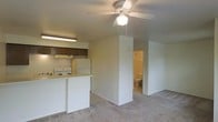 This single-floor unit offers a practical, open-concept layout that seamlessly connects the kitchen and living area. The kitchen is equipped with a white electric range, a refrigerator, and a peninsula-style counter that provides both workspace and a natural division between the cooking and living zones. Dark-toned upper cabinetry with under-cabinet lighting adds contrast against the lighter walls and surfaces, while a ventilation hood is positioned above the range. The living area features neutral wall-to-wall carpeting that extends throughout, contributing to a cohesive and understated aesthetic. A ceiling fan with an integrated light fixture is centrally positioned overhead, offering both ventilation and ambient lighting. The layout includes a hallway that leads to a bathroom, which is partially visible and appears to include standard fixtures. The overall design is clean and functional, with neutral tones dominating the color palette. The open flow between the kitchen and living space maximizes the usable area, making the layout efficient and easy to navigate.