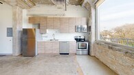 Located on the fourth floor at 714 Walbridge St in Kalamazoo, Michigan, this industrial-style loft unit offers a compelling blend of raw architectural character and modern conveniences. The open-plan living area is defined by exposed original brick walls, polished concrete floors, and a dramatic exposed concrete ceiling with visible structural columns and piping — hallmarks of adaptive reuse design. An abundance of natural light pours in through a series of large windows that line the exterior walls, framing treetop views of the surrounding Kalamazoo skyline. A glass door leads directly to a private balcony, further enhancing the connection to the outdoors. The kitchen is thoughtfully equipped with light wood cabinetry, white countertops, and a full suite of stainless steel appliances including a refrigerator, electric range, over-the-range microwave, and dishwasher. A separate bedroom area is partitioned from the main living space, featuring clean white walls, a wall-mounted mini-split HVAC unit, and access to a bathroom. The entryway is compact and functional, with a modern front door and ample room for transitioning into the open living space. The unit's industrial aesthetic is balanced by contemporary finishes, creating a distinctive and characterful living environment.