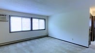 This single-floor space offers a straightforward and functional layout designed for comfortable everyday living. The interior is finished with wall-to-wall carpet throughout, providing a soft and warm underfoot feel that contributes to a cozy atmosphere. The main living area is generously sized, featuring large windows fitted with horizontal blinds that allow abundant natural light to fill the room while maintaining privacy. The white walls provide a neutral, versatile backdrop that adapts well to a variety of décor styles. A textured popcorn ceiling adds a classic residential character to the space. Baseboard heating runs along the perimeter of the room, offering an efficient and consistent heat source during colder months. The open wall space provides ample flexibility for furniture arrangement. Overall, the space presents a clean, well-maintained environment with practical finishes and a bright, airy ambiance suited for a range of occupants looking for a move-in ready setting.