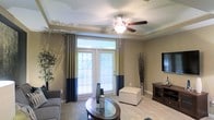 This single-floor residence offers a thoughtfully designed living space that balances comfort and style. The living area serves as the centerpiece of the home, showcasing elegant crown molding that frames the ceiling and adds a refined architectural touch. A classic ceiling fan with a built-in light fixture provides both function and a sense of warmth to the room.

Large French doors dominate one wall, fitted with horizontal blinds and dressed with eye-catching two-tone curtains in navy blue and yellow-green, allowing an abundance of natural light to flood the interior while offering a view of the lush greenery outside. The neutral, warm-toned walls create a versatile backdrop that complements the space's decor.

The living area is carpeted, adding a soft, comfortable underfoot feel, and is furnished with a sleek glass-topped coffee table, a plush sofa, and a padded ottoman that doubles as additional seating. A dark wood media console anchors the opposite wall, providing practical storage and display space. Decorative elements, including tall vases with branches, add an organic, artistic flair. Overall, the space exudes a clean, welcoming ambiance ideal for both relaxing and entertaining.