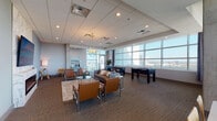 This contemporary amenity lounge is thoughtfully designed to offer both relaxation and entertainment in an elevated, high-rise setting. Floor-to-ceiling windows span multiple walls, flooding the space with abundant natural light and framing sweeping panoramic city views that serve as a dramatic backdrop throughout.

The main lounge area is anchored by a sleek, wall-mounted electric fireplace set against a marble surround, complemented by a large flat-screen television. Comfortable seating arrangements feature tan leather chairs with chrome frames and a plush sofa, centered around glass-top accent tables and area rugs. An elegant globe chandelier adds a refined decorative touch overhead, while recessed lighting provides consistent illumination across the carpeted floors.

A dedicated game area includes a foosball table and a shuffleboard table, offering casual entertainment options. Exposed concrete columns add an industrial architectural contrast to the otherwise polished, modern interior.

A separate, windowed meeting or dining nook provides a more intimate setting, furnished with a white table and natural wood chairs, bathed in daylight from a large corner window with expansive skyline views. An adjacent lounge alcove with mid-century-style wooden chairs and curated wall art offers a quieter retreat. Together, these distinct zones make this space a versatile and well-appointed communal amenity.