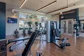 Located at 2007 Monterey Circle in Alameda, California, this fitness center offers a thoughtfully designed workout space spread across a single open floor. The room features soaring ceilings with exposed ductwork, lending an industrial-modern aesthetic complemented by clean white walls and a bold teal accent wall. Large multi-pane windows line the perimeter, flooding the interior with natural light and providing views of the surrounding greenery and outdoor amenities.

The flooring throughout is a warm-toned hardwood, providing both durability and visual appeal. The cardio zone is well-appointed with multiple treadmills, elliptical machines, and rowing ergometers, each equipped with personal display screens. A wall-mounted flat-screen TV serves this area for entertainment during workouts.

The strength training section features a full rack of dumbbells in varying weights, adjustable benches, a cable machine, and a range of functional training tools including medicine balls and resistance equipment. A large floor-to-ceiling mirror spans one wall, enhancing the sense of space while allowing users to monitor their form. Pendant and linear lighting fixtures supplement the natural light for evening use. The facility is accessed through glass double doors and presents a clean, professional environment suited for a comprehensive fitness routine.