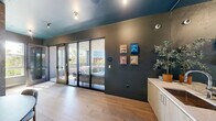 Located at 600 St Albans Dr in Raleigh, NC, this upscale amenity lounge is a compact yet thoughtfully designed communal space that seamlessly blends interior comfort with outdoor living. The interior is anchored by wide-plank light oak hardwood flooring, complemented by a striking teal-painted ceiling that adds a bold design accent. Dark, textured wall treatments — including an ornate botanical-patterned wallcovering — lend an air of contemporary sophistication throughout. A long run of light wood cabinetry with a white stone countertop and integrated sink provides a functional wet bar area, topped by a large wall-mounted display screen above. A rounded dining table with a velvet green banquette tucks neatly against the feature wall, creating an intimate seating nook. Full-height black-framed glass sliding and folding doors connect the interior directly to a covered outdoor terrace. The terrace is generously furnished with plush sectional sofas, accent chairs, and low cocktail tables, all set on a grey wood deck beneath a paneled ceiling with pendant lighting. Open railings frame sweeping views of the surrounding Raleigh skyline and lush tree canopy, making the outdoor space ideal for both relaxed gatherings and social events.