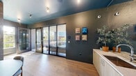 Located at 600 St Albans Dr in Raleigh, NC, this upscale amenity lounge is a compact yet thoughtfully designed communal space that seamlessly blends interior comfort with outdoor living. The interior is anchored by wide-plank light oak hardwood flooring, complemented by a striking teal-painted ceiling that adds a bold design accent. Dark, textured wall treatments — including an ornate botanical-patterned wallcovering — lend an air of contemporary sophistication throughout. A long run of light wood cabinetry with a white stone countertop and integrated sink provides a functional wet bar area, topped by a large wall-mounted display screen above. A rounded dining table with a velvet green banquette tucks neatly against the feature wall, creating an intimate seating nook. Full-height black-framed glass sliding and folding doors connect the interior directly to a covered outdoor terrace. The terrace is generously furnished with plush sectional sofas, accent chairs, and low cocktail tables, all set on a grey wood deck beneath a paneled ceiling with pendant lighting. Open railings frame sweeping views of the surrounding Raleigh skyline and lush tree canopy, making the outdoor space ideal for both relaxed gatherings and social events.