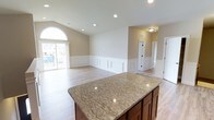 This well-appointed single-story home offers three bedrooms and two bathrooms across a generously sized floor plan. The open-concept living and kitchen area serves as the heart of the home, highlighted by a large kitchen island topped with striking granite countertops and warm wood cabinetry beneath. The living area flows seamlessly from the kitchen, featuring classic wainscoting along the lower walls that adds a refined architectural detail throughout the space. Light-toned hardwood-style flooring runs continuously across the main living areas, contributing to a bright and cohesive aesthetic. A notable arched window in the living area invites abundant natural light, further enhancing the open and airy atmosphere. Recessed lighting throughout ensures the space is well-lit at all hours. Vaulted ceilings in the living area lend a sense of volume and grandeur. Neutral wall tones create a versatile backdrop well-suited to a variety of interior styles. Multiple doorways lead to additional rooms, including the bedrooms and bathrooms, offering a practical and functional layout. Overall, this home combines classic design elements with modern finishes to create a comfortable and inviting living environment.