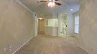 This single-story space is designed with an open-concept layout that connects the main living area to the kitchen, creating a functional and cohesive floor plan. The living area is finished with wall-to-wall carpeting in a neutral beige tone, providing a comfortable and understated foundation. Crown molding runs along the upper perimeter of the living space, adding a subtle architectural detail. A ceiling fan with a light fixture is centrally positioned, offering both ambient lighting and air circulation.

The kitchen is partially separated from the living area by a half-wall pass-through, which also serves as a breakfast bar or counter space. White cabinetry lines the kitchen walls, complemented by white appliances including a refrigerator and a range, maintaining a clean and uniform aesthetic. Recessed lighting in the kitchen ensures adequate task lighting. A hallway visible from the main living area leads to additional rooms, suggesting a straightforward and accessible layout. Natural light filters into the space through a window, contributing to a bright, open atmosphere throughout. The overall palette of soft greens and neutrals gives the space a calm, move-in-ready feel.