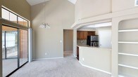 This single-floor space is defined by its dramatic vaulted ceilings, which rise to an impressive height in the main living area, creating a sense of openness and grandeur. A modest chandelier hangs centrally, adding a refined touch to the airy atmosphere. The living area is finished with plush neutral carpeting that extends throughout, contributing to a warm and cohesive aesthetic.

The open-concept layout connects the living space seamlessly to the kitchen, which is elevated slightly and separated by a curved breakfast bar with a light stone countertop — an ideal surface for casual dining or entertaining. The kitchen is well-appointed with warm medium-toned wood cabinetry, a decorative upper wine rack, subway tile backsplash, and stainless steel appliances including a refrigerator and sink. A tray ceiling detail frames the kitchen area, adding architectural interest.

Neutral beige tones are carried throughout the walls, lending a calm and inviting character to the space. A hallway visible from the main living area provides access to additional rooms. Overall, the space balances functional design with architectural character.