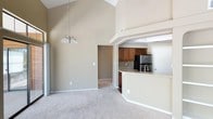 This single-floor space is defined by its dramatic vaulted ceilings, which rise to an impressive height in the main living area, creating a sense of openness and grandeur. A modest chandelier hangs centrally, adding a refined touch to the airy atmosphere. The living area is finished with plush neutral carpeting that extends throughout, contributing to a warm and cohesive aesthetic.

The open-concept layout connects the living space seamlessly to the kitchen, which is elevated slightly and separated by a curved breakfast bar with a light stone countertop — an ideal surface for casual dining or entertaining. The kitchen is well-appointed with warm medium-toned wood cabinetry, a decorative upper wine rack, subway tile backsplash, and stainless steel appliances including a refrigerator and sink. A tray ceiling detail frames the kitchen area, adding architectural interest.

Neutral beige tones are carried throughout the walls, lending a calm and inviting character to the space. A hallway visible from the main living area provides access to additional rooms. Overall, the space balances functional design with architectural character.