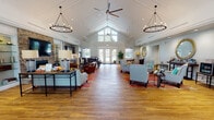 Located at 3258 Turtle Point Dr in Fayetteville, NC, this single-story clubhouse is designed to impress with its dramatic vaulted, cathedral-style ceilings that soar overhead throughout the main gathering area. Warm hardwood flooring flows seamlessly across the open-concept layout, anchoring the space with a sense of continuity and elegance. Statement ring chandeliers and large ceiling fans are suspended from the high peaks, complementing the abundant natural light that pours in through oversized windows and French doors leading to the outdoors.

The central living area serves as the social heart of the space, featuring a striking floor-to-ceiling stacked stone fireplace flanked by built-in glass shelving and a mounted flat-screen television. Comfortable seating arrangements, including plush sofas and leather armchairs, are thoughtfully positioned around vibrant area rugs to create intimate conversation zones within the expansive room.

A dedicated reception and office area near the entrance offers a professional workspace with a large desk, seating for guests, wall-mounted display, and direct access to the exterior. A secondary lounge area provides additional seating clusters with upholstered armchairs, a decorative console, and a circular mirror as a focal point. Throughout, the design balances an inviting, residential warmth with polished, community-oriented functionality.