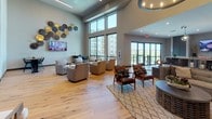 This thoughtfully designed single-floor clubhouse space blends rustic charm with modern sophistication throughout its open, airy layout. The entryway immediately impresses with dramatic soaring ceilings and striking reclaimed wood plank accent walls that set a warm, textured tone. Light natural hardwood flooring flows seamlessly throughout, complementing the neutral color palette and curated furnishings.

The main lounge area is a standout feature, bathed in natural light from floor-to-ceiling windows that frame panoramic views of the surrounding landscape and neighboring buildings. Comfortable seating arrangements — including leather lounge chairs and upholstered sofas — are thoughtfully positioned around a mounted flat-screen television and anchored by a woven area rug. A decorative wall display of metallic disc art adds an artistic focal point.

The kitchen and bar area is well-appointed with charcoal gray shaker-style cabinetry, white quartz countertops, a large center island with bar seating, and dual wall-mounted televisions backed by a tiled accent wall. Ample storage is provided through full-height cabinetry, and a beverage station adds functional convenience.

A more intimate seating nook near the entry features a rustic reclaimed wood console table, accent chairs, and Western-inspired décor including a longhorn mount, creating a welcoming and characterful atmosphere throughout this versatile communal space.