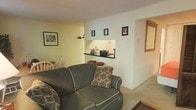 This single-floor space offers a comfortable and functional layout with a warm, understated aesthetic. The living area serves as the central gathering space, furnished with a plush sofa accented by decorative throw pillows and neutral carpeting underfoot. Light-colored walls contribute to an airy feel throughout.

A distinctive built-in recessed shelf along one wall serves as a focal display area, illuminated to highlight decorative items and personal accents. Framed artwork and photographs are arranged along the walls, adding a personalized touch to the space.

An open pass-through or nook connects the living area to what appears to be a small bar or counter area, offering a sense of flow between spaces. To one side, a dining area is partially visible, featuring wooden seating that complements the overall neutral color palette.

A framed mirror or artwork in a warm wood tone is visible near a hallway or adjoining room, suggesting additional living areas beyond. The consistent use of soft, muted tones throughout—from the walls to the flooring—creates a cohesive and inviting atmosphere well-suited for everyday living.