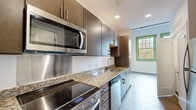 This thoughtfully designed single-floor unit blends modern finishes with a clean, functional layout. The galley-style kitchen is a standout feature, showcasing rich dark wood cabinetry, granite countertops with a stainless steel backsplash, and a full suite of stainless steel appliances including a smooth-top electric range, over-the-range microwave, dishwasher, and a full-size refrigerator. Recessed lighting illuminates the space, while a distinctive green-trimmed window at the end of the kitchen draws in natural light and adds a charming architectural accent. The open living area flows seamlessly from the kitchen, featuring wide-plank light hardwood flooring and bright white walls that create an airy, welcoming atmosphere. The bedroom is particularly impressive, with soaring ceilings, the same warm hardwood floors, and a large green-trimmed window that floods the room with natural light. The bathroom is finished in a crisp, contemporary style with large-format tile flooring, a dark wood vanity with a white countertop, a wall-mounted mirror, and a fully tiled walk-in shower enclosed by a glass door with brushed nickel hardware throughout. Together, these spaces offer a cohesive, modern aesthetic with high-quality finishes at every turn.