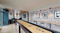 This thoughtfully designed amenity lounge offers a seamless blend of recreation, comfort, and contemporary style across a single open floor. The space is anchored by warm wood-paneled ceilings that run throughout, lending a natural warmth that contrasts beautifully with the light hardwood flooring underfoot.

The recreation area features a full-length shuffleboard table as a centerpiece, complemented by a dedicated lounge zone with a generously sized sectional sofa accented with teal throw pillows. Decorative laser-cut screen panels filter natural light from the windows, casting delicate patterns throughout the space and adding an artistic architectural element.

A media wall clad in textured tile surrounds a large flat-screen TV, with sleek lounge chairs arranged for comfortable viewing. Adjacent to this area, a quieter sitting nook provides a more intimate retreat.

The dining and social area features bar-height communal tables with pendant lighting above, ideal for gathering. The kitchenette is outfitted with light wood cabinetry, a stainless steel wall oven, ample counter space, and a patterned tile backsplash that adds visual interest.

Throughout the lounge, curated artwork adorns the walls, and the open layout encourages both lively interaction and relaxed downtime, making it a well-rounded and inviting communal destination.