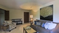 This single-floor space offers a thoughtfully arranged living area that combines comfort and style. The living room serves as the centerpiece, featuring soft wall-to-wall carpeting that adds warmth underfoot and contributes to the room's cozy ambiance. A gray upholstered sofa, accented with decorative throw pillows in shades of blue and white, anchors the seating area alongside a patterned accent chair, creating a welcoming arrangement ideal for relaxing or entertaining. A rectangular coffee table with a decorative bowl centerpiece sits at the heart of the space, while a slim console table along the wall provides additional surface space. Warm ambient lighting is provided by a classic table lamp with a cream shade, casting a soft glow throughout the room. A large nature-inspired wall art piece featuring lush foliage adds a vivid focal point and a touch of the outdoors to the interior. The space is completed by a solid wood entry door, offering a clear sense of enclosure and privacy. The neutral wall tones and ceiling complement the overall palette, creating a harmonious and balanced environment that feels both functional and aesthetically pleasing.