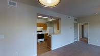 This single-story space is designed with a functional, open layout that connects the main living area to the kitchen. The living room is spacious, featuring wall-to-wall carpeting in a neutral gray tone that extends throughout the space. A ceiling fan is centrally mounted, providing both light and airflow, while a pendant light fixture over the dining area adds a decorative touch. A window along one wall allows natural light to filter in, brightening the room. Neutral-toned walls keep the space feeling clean and versatile.

The kitchen is accessible via a pass-through opening that also serves as a breakfast bar, creating a seamless connection between cooking and living spaces. Inside, the kitchen features warm honey-toned wood cabinetry, a white refrigerator, and an electric range, all set on hardwood-style flooring that contrasts with the carpeting in the adjacent rooms. Under-cabinet lighting provides functional illumination. Built-in shelving is visible near the kitchen area, offering convenient storage. A doorway visible from the living room leads further into the unit, suggesting additional private rooms beyond.