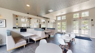 This well-appointed single-floor community clubhouse offers a diverse range of amenities across several distinct, thoughtfully designed zones. The great room is an architectural highlight, boasting soaring vaulted ceilings with exposed red wood trusses, an abundance of natural light streaming through tall clerestory and double-hung windows, warm hardwood flooring, and a stone accent wall. Comfortable lounge seating, curated artwork, and vibrant décor accents create an inviting, residential atmosphere. Adjacent to the great room, a bar-height counter with black granite tops and industrial-style metal stools runs along a window-lined wall, providing an ideal space for casual work or socializing. The café and dining area features a textured ceiling, geometric pendant lighting, upholstered booth seating, and modern round tables with contemporary chairs, all set on large-format slate tile flooring. Multiple sets of French doors open to outdoor views, flooding the space with natural light. A café kitchen includes a white subway tile backsplash, dark countertops, stainless steel appliances, and a mounted flat-screen TV. The fitness center is equipped with cardio machines, a power rack, free weights, and floor-to-ceiling mirrors, offering a comprehensive workout facility. Throughout, a cohesive palette of soft blues, whites, and grays ties the space together.
