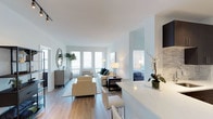 This two-bedroom Quincy residence is designed with a modern, open-concept layout that promotes a seamless flow between living and dining areas. The interior is bathed in natural light, thanks to multiple windows fitted with horizontal blinds that allow for adjustable brightness throughout the day. Crisp white walls and smooth ceilings enhance the sense of openness, while light hardwood flooring runs consistently through the main living spaces, adding warmth and cohesion to the overall design.

The living area is generously proportioned, comfortably accommodating a seating arrangement with cream-toned upholstered chairs and a sofa accented with teal throw pillows. A sleek, dark coffee table grounds the space, complemented by coordinated floor and table lamps that provide layered ambient lighting. A decorative oval mirror and greenery add character to the room.

The kitchen features a white countertop peninsula that doubles as a breakfast bar with seating, offering both functionality and a natural partition from the living space. A glimpse into the bedroom reveals a well-appointed space with soft blue bedding, continuing the unit's cool, calming color palette throughout.