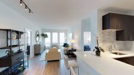 This two-bedroom Quincy residence is designed with a modern, open-concept layout that promotes a seamless flow between living and dining areas. The interior is bathed in natural light, thanks to multiple windows fitted with horizontal blinds that allow for adjustable brightness throughout the day. Crisp white walls and smooth ceilings enhance the sense of openness, while light hardwood flooring runs consistently through the main living spaces, adding warmth and cohesion to the overall design.

The living area is generously proportioned, comfortably accommodating a seating arrangement with cream-toned upholstered chairs and a sofa accented with teal throw pillows. A sleek, dark coffee table grounds the space, complemented by coordinated floor and table lamps that provide layered ambient lighting. A decorative oval mirror and greenery add character to the room.

The kitchen features a white countertop peninsula that doubles as a breakfast bar with seating, offering both functionality and a natural partition from the living space. A glimpse into the bedroom reveals a well-appointed space with soft blue bedding, continuing the unit's cool, calming color palette throughout.