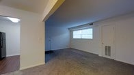 This single-floor residential space offers a clean, functional layout designed for comfortable everyday living. The main living area is finished with wall-to-wall carpet in a neutral tone, providing a warm and understated foundation throughout. White walls and a smooth ceiling create a bright, open atmosphere, while a floor-level HVAC vent indicates a central climate control system integrated into the space.

A defining architectural feature is the structural beam that runs along the ceiling, creating a subtle visual division between the main living area and an adjoining room. This secondary carpeted space receives natural light through a large window fitted with blinds, giving it an airy feel.

The layout includes a hallway visible from the main living area, connecting to additional rooms via interior doors. The transition from carpet to hard flooring is noticeable in the hallway zone, suggesting a practical separation between living and utility areas. The doors throughout are standard white panel style, maintaining a consistent and simple aesthetic.

Overall, the space presents a no-frills, move-in-ready environment with a neutral palette, adequate natural light, and a sensible floor plan suited for a variety of living arrangements. The straightforward design makes it easy to personalize and furnish to individual preference.