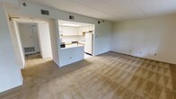 This single-floor residence offers a practical and functional layout centered around an open-concept living and kitchen area. The spacious living room is finished with neutral-toned wall-to-wall carpeting, creating a warm and understated atmosphere throughout the main space. The kitchen is positioned as a semi-open peninsula layout, separated from the living area by a raised breakfast bar counter. It features white cabinetry above and below the counter, a white refrigerator, a range with an oven, and a dishwasher, all set against a tile floor. A recessed overhead light panel provides bright, even illumination in the kitchen. The white walls and light-colored finishes throughout the space contribute to a clean, bright aesthetic. Ventilation is provided via ceiling-mounted HVAC vents visible in both the living and kitchen areas. A hallway visible to the left of the kitchen suggests access to additional rooms such as a bedroom or bathroom. The overall design is modest and efficient, making practical use of the available floor space while maintaining an open, airy feel. This residence is well-suited for those seeking a simple, move-in-ready living environment with a connected kitchen and living space.