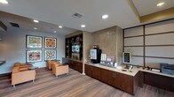 Located at 12325 Shadow Creek Pkwy in Pearland, TX, this single-floor hospitality space is thoughtfully designed to offer guests a comfortable and functional environment. The grand entrance foyer immediately sets the tone with its soaring ceilings, large glass doors, and striking geometric-patterned tile flooring that reflects an abundance of natural light. Lush potted plants flank the entryway, adding a fresh, welcoming ambiance.

The lounge area features warm orange leather seating arranged around small circular tables, creating an inviting space for relaxation or casual conversation. A bold gallery wall displays four large, colorful framed artworks with geometric and abstract patterns, adding a vibrant focal point to the room. Dark wood built-in shelving with a mounted display screen lines one wall, complementing the rich hardwood-style flooring.

Adjacent to the lounge, a self-serve beverage station offers a coffee machine, cup dispensers, and a variety of amenities set along a continuous dark wood cabinetry counter with a stone-tile accent wall behind it. A dedicated business center provides two workstations with desktop computers, set against a striking grid-panel wall feature, with natural light filtering in through draped windows. The space as a whole maintains a cohesive, modern aesthetic through consistent warm tones, recessed lighting, and refined finishes throughout.