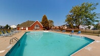 Located at 1040 Windward Dr in London, Ohio, this outdoor community pool facility offers a well-appointed recreational space designed for warm-weather enjoyment. The centerpiece is a generously sized rectangular swimming pool with clearly marked depth zones, including a designated shallow water area, and equipped with multiple entry points via pool ladders. The water is maintained to a clear, inviting turquoise appearance.

Surrounding the pool is a spacious concrete deck furnished with numerous blue lounge chairs and seating arrangements, providing ample space for sunbathing and relaxation on all sides. The perimeter of the pool area is enclosed by a black wrought iron fence, ensuring a safe and defined boundary.

Adjacent to the pool is a single-story red brick clubhouse building featuring a distinctive A-frame gabled roofline with a large arched window, adding architectural character to the facility. The clubhouse serves as a support structure for the pool area.

The grounds are complemented by mature trees that provide natural shade along the fencing perimeter, and the open setting offers views of the surrounding neighborhood and the London city water tower in the background. The overall environment is open, bright, and well-suited for community recreation under clear skies.