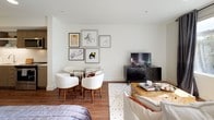 This single-floor residence offers a thoughtfully designed open-concept layout that seamlessly connects the living, dining, and sleeping areas. Warm hardwood flooring runs throughout, providing a cohesive foundation that ties each zone together.

The living area is anchored by a large window dressed with floor-to-ceiling charcoal drapes, allowing natural light to filter in while framing views of the surrounding neighborhood. A plush white sofa, rustic wood coffee table, and a sleek black media console furnish the space with a balanced mix of modern and organic elements.

The dining area sits adjacent to the living space, featuring a glass-top table surrounded by sculpted white chairs beneath a curated gallery wall of framed artwork. The galley-style kitchen is outfitted with flat-front wood-toned cabinetry, white countertops, stainless steel appliances — including a refrigerator, range, and built-in microwave — and a textured tile backsplash. A compact breakfast bar with stools adds functional versatility near the entryway.

The bedroom accommodates a full-sized bed alongside a closet and offers enough space for additional furnishings. The bathroom features a tub-shower combination, wood-toned vanity with an integrated sink, large mirror, and crisp white walls, maintaining the clean aesthetic found throughout the residence.