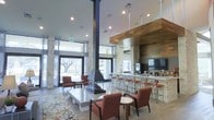 This contemporary single-floor gathering space blends rustic and modern design elements to create a welcoming, upscale atmosphere. At the heart of the room stands a dramatic suspended angular fireplace with a sleek black flue, serving as a bold architectural centerpiece that anchors the open-plan layout. Surrounding the fireplace, comfortable seating arrangements feature warm terracotta-toned upholstered chairs and patterned area rugs, creating an inviting lounging environment.

Adjacent to the seating area, a full-service bar runs along one wall, finished with a natural stone facade and topped with a wood-paneled accent soffit above. Bar-height seating lines the counter, complemented by a large flat-screen television mounted overhead — ideal for social gatherings or events.

The space benefits from soaring ceilings with recessed lighting and an abundance of natural light pouring in through expansive floor-to-ceiling windows and glass doors. The windows frame views of mature trees outside, further connecting the interior to its natural surroundings. Structural columns clad in natural limestone add texture and complement the mix of wood, stone, and neutral tones throughout. The light hardwood-style flooring ties all the design elements together, reinforcing the space's modern yet warm aesthetic.