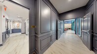 This elegantly designed single-floor space makes a striking first impression through a corridor clad in raised-panel gray wainscoting, gold-accented door hardware, and polished marble-look tile flooring that carries throughout the entire layout. The corridor leads into a dramatic main lounge area distinguished by soaring double-height ceilings and a bold, illuminated gold ceiling feature that draws the eye upward. A sleek, glass-enclosed linear fireplace serves as a central architectural focal point, flanked by a built-in bench and a wall-mounted flat-screen television set into a paneled gray accent wall. The lounge is furnished with a teal sectional sofa, vibrant orange accent chairs, patterned area rugs, and carefully curated decorative accessories, creating a warm yet contemporary ambiance. Floor-to-ceiling windows dressed in teal drapes flood the space with natural light and provide views of the surrounding exterior. Transitioning from the main lounge, a more intimate dining and café-style area features rich teal walls adorned with bold framed artwork, plush channeled terracotta banquette seating, round wooden tables, and warm globe pendant lighting, offering a cozy and inviting retreat. Together, the spaces combine refined architectural detailing with a vibrant, curated color palette to deliver an atmosphere that is both sophisticated and welcoming.