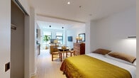 This single-floor studio unit offers a well-organized open-concept layout that seamlessly integrates sleeping, living, and kitchen areas into one cohesive space. Light oak hardwood floors run throughout, lending warmth and continuity across every zone.

The sleeping area is anchored by a full bed dressed in rich earth tones, accompanied by a wooden dresser with a mirror and a small round dining table with woven-seat chairs — a versatile touch that maximizes functionality. Framed wall art adds a refined, curated quality to the space.

The living area features a neutral linen sofa paired with a tufted blue ottoman, a wall-mounted flat-screen TV, and a wooden work table positioned beneath a pair of casement windows. Natural light filters in generously, creating a bright and airy atmosphere. A fiddle-leaf fig plant contributes a lively, organic element.

The kitchen is compact yet fully equipped, showcasing a striking contrast of matte black upper cabinetry and warm wood lower cabinets. Stainless steel appliances — including a refrigerator, gas range, microwave, and dishwasher — are neatly integrated alongside white stone countertops. The overall design is clean, modern, and highly functional, making excellent use of every square foot in this intelligently planned studio.