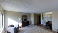 This single-floor space centers around a generously sized bedroom that combines comfort and functionality. The room features plush carpeting underfoot and a textured popcorn ceiling overhead. Natural light enters through unseen windows, casting soft shadows across the neutral-toned walls and illuminating the space with a calm, understated ambiance. A dark wood dresser with an ornate framed mirror serves as a focal point along one wall, accompanied by a classic floor lamp and a leather accent chair, adding character to the space. The bedroom transitions into a hallway finished with hardwood flooring, which leads to what appears to be a pair of closets, offering generous storage. Adjacent to the hallway, a secondary area is visible, featuring white cabinetry that suggests a kitchenette or utility space with ample cabinet storage. The overall layout is practical and cohesive, with a clear distinction between the carpeted sleeping area and the hard-floored utility zones. The neutral color palette throughout creates a unified, livable environment suited for everyday comfort.