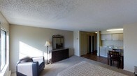 This single-floor space centers around a generously sized bedroom that combines comfort and functionality. The room features plush carpeting underfoot and a textured popcorn ceiling overhead. Natural light enters through unseen windows, casting soft shadows across the neutral-toned walls and illuminating the space with a calm, understated ambiance. A dark wood dresser with an ornate framed mirror serves as a focal point along one wall, accompanied by a classic floor lamp and a leather accent chair, adding character to the space. The bedroom transitions into a hallway finished with hardwood flooring, which leads to what appears to be a pair of closets, offering generous storage. Adjacent to the hallway, a secondary area is visible, featuring white cabinetry that suggests a kitchenette or utility space with ample cabinet storage. The overall layout is practical and cohesive, with a clear distinction between the carpeted sleeping area and the hard-floored utility zones. The neutral color palette throughout creates a unified, livable environment suited for everyday comfort.