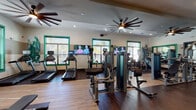 This fitness center is a thoughtfully designed, single-floor facility that provides a comprehensive workout environment. The space is defined by its warm wood-look flooring, neutral two-tone walls accented with bold green window trim, and recessed lighting complemented by striking multi-blade ceiling fans that enhance air circulation throughout.

The cardio zone features a row of Matrix treadmills and elliptical machines, each equipped with individual digital consoles for personalized training. Wall-mounted flat-screen TVs are positioned along the cardio row, offering entertainment during workouts. Natural light filters in through multiple green-trimmed windows fitted with blinds, creating a bright and energizing atmosphere.

The strength training area is well-stocked with Matrix cable and resistance machines arranged to maximize floor space and ease of movement. A dedicated free weight section includes a full rack of dumbbells displayed against floor-to-ceiling mirrors, which amplify the sense of space and allow users to monitor their form. The mirrored walls extend throughout much of the facility, contributing to the open, expansive feel of the room.

A separate adjoining room, visible through interior windows and glass doors with green framing, appears to offer additional fitness or recreational amenities. Overall, the fitness center is clean, modern, and functionally organized for a well-rounded exercise experience.