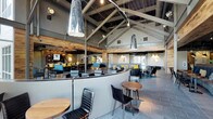 This single-story space showcases a striking vaulted ceiling with exposed dark wood beams and steel structural elements, creating an open, airy atmosphere with considerable vertical volume. The industrial-chic aesthetic is complemented by reclaimed wood accents along the upper walls, adding warmth to the otherwise contemporary palette of grey and steel tones.

The main dining area features a curved banquette booth as a central design element, upholstered in deep charcoal and anchored by round wooden bistro tables with sleek black chairs. Large pendant lights—sculptural and elongated—hang dramatically from the ceiling, serving as focal points throughout the space. Pops of teal and yellow in the seating and décor provide a lively, modern contrast.

Toward the rear, a lounge area offers a more relaxed arrangement with softer seating, while a bar area is visible in the background. Large windows along the perimeter allow natural light to filter in, brightening the space. The slate-grey tile flooring ties all zones together cohesively, reinforcing the space's polished yet approachable character.