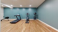 Located at 113 N Ohio Ave in Sidney, Ohio, this fitness room offers a well-appointed exercise space designed for efficient, focused workouts. The room features warm hardwood flooring throughout, providing a durable and attractive surface suited for a variety of exercise activities. Crisp white walls are complemented by a bold teal accent wall, lending the space a fresh, energizing atmosphere. Recessed ceiling lighting ensures even, bright illumination across the entire floor area. A prominently routed teal HVAC duct runs along the upper wall, providing climate control and maintaining air circulation during workouts. The space is equipped with a range of cardio and strength equipment, including a treadmill, elliptical trainer, rowing machine, stationary bike, adjustable weight bench, dumbbell rack, cable machine, and a stability ball station — offering a comprehensive workout experience. Clean white baseboards frame the room, and an accessible exit door with a lit exit sign ensures safety compliance. The overall design is minimal and functional, with a thoughtful color palette and practical layout that maximizes usable floor space while maintaining an inviting, motivating environment for fitness.