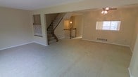This multi-level home spans three floors, offering generous living space throughout. The main level showcases a large, open living area with plush wall-to-wall carpeting that creates a warm and comfortable atmosphere. Neutral, light-toned walls throughout the space lend a clean and versatile backdrop, ready to accommodate a variety of interior design styles.

A central staircase serves as a key architectural feature, finished with classic white balusters and a wood handrail, connecting all three floors with a timeless, traditional aesthetic. The open stairwell design allows sightlines across multiple levels, enhancing the sense of openness and flow throughout the home.

Natural light filters into the living area through well-placed windows, brightening the neutral interior and creating an airy feel. A ceiling fan with a light fixture is installed in the living area, adding both functionality and a classic design touch.

Baseboard heating units are visible along the walls, indicating a reliable heating system integrated throughout the home. The open-concept layout of the main living area allows for flexible furniture arrangement and entertaining space. Overall, this three-floor home combines practical design with comfortable, neutral finishes, offering a spacious and adaptable living environment across all levels.