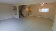 This multi-level home spans three floors, offering generous living space throughout. The main level showcases a large, open living area with plush wall-to-wall carpeting that creates a warm and comfortable atmosphere. Neutral, light-toned walls throughout the space lend a clean and versatile backdrop, ready to accommodate a variety of interior design styles.

A central staircase serves as a key architectural feature, finished with classic white balusters and a wood handrail, connecting all three floors with a timeless, traditional aesthetic. The open stairwell design allows sightlines across multiple levels, enhancing the sense of openness and flow throughout the home.

Natural light filters into the living area through well-placed windows, brightening the neutral interior and creating an airy feel. A ceiling fan with a light fixture is installed in the living area, adding both functionality and a classic design touch.

Baseboard heating units are visible along the walls, indicating a reliable heating system integrated throughout the home. The open-concept layout of the main living area allows for flexible furniture arrangement and entertaining space. Overall, this three-floor home combines practical design with comfortable, neutral finishes, offering a spacious and adaptable living environment across all levels.
