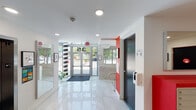 Located at 35 SW Sixth Ave in Miami, FL, this ground-floor space encompasses a clean, contemporary lobby and a fully equipped gym. The entrance greets visitors with floor-to-ceiling glass doors framed in aluminum, polished tile flooring, and a bold black-and-white striped column that adds a striking design accent. Recessed lighting illuminates the bright, open lobby, which features crisp white walls accented with vibrant red trim — a color theme carried consistently throughout the space.

Adjacent to the main entrance is a well-organized mailroom, outfitted with rows of individual resident mailboxes topped with vivid red countertops. Large mirrors and framed artwork contribute to the tidy, functional aesthetic of the area. Clearly marked stairwell and elevator access points provide easy navigation to upper floors.

The on-site fitness center offers a comprehensive workout environment. The cardio area features multiple treadmills and a stationary bike positioned in front of a floor-to-ceiling glass wall, providing natural light and outdoor views of the surrounding greenery. The strength training section includes a multi-function cable machine, leg extension equipment, and free weights, all set on warm wood-tone flooring. Exposed ductwork on the ceiling lends an industrial character to the gym, complementing its practical, fully functional design.