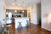 Located at 712 Promenade Place in Virginia Beach, Virginia, this single-floor residence offers a well-designed layout with contemporary finishes throughout. The kitchen is a standout feature, showcasing gray shaker-style cabinetry, granite countertops, a mosaic tile backsplash, stainless steel appliances, and a large island with bar seating and pendant lighting overhead — ideal for both cooking and casual dining. Wood-style flooring runs through the main living areas, adding warmth and visual continuity. The living room is bright and inviting, centered around a sliding glass door that opens to a private balcony with outdoor views. Multiple bedrooms are carpeted for comfort; one features dual windows that fill the room with natural light, while another includes direct access to an ensuite bathroom. The bathrooms are clean and functional — one offers a full soaking tub with a shower combination, while the other provides a walk-in shower stall. Both feature neutral tile flooring and chrome fixtures. A dedicated laundry room houses a full-size washer and dryer with overhead shelving for added storage. The overall design palette of soft neutrals and warm tones creates a cohesive, move-in-ready atmosphere throughout the space.