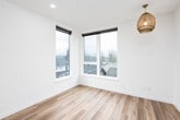 This contemporary single-floor unit showcases a clean, minimalist design with crisp white walls and light hardwood-style flooring that flows consistently throughout the space. The compact kitchenette is thoughtfully appointed with warm wood cabinetry, a white quartz countertop, a matte black sink faucet, open floating shelves, and built-in appliances including a refrigerator and microwave — all neatly integrated into a streamlined layout. Adjacent to the kitchen, the main living area benefits from an abundance of natural light, courtesy of large corner windows that frame views of the surrounding neighborhood and tree line. A decorative woven pendant light fixture adds a touch of warmth and texture to the otherwise neutral palette. The bathroom features matching light hardwood-style flooring, a wood-tone vanity with a white countertop, a toilet, and matte black hardware accents, maintaining the modern aesthetic carried throughout the unit. A dedicated electrical panel is conveniently accessible within the unit. Additional space is provided by a separate, versatile room with clean white walls, suitable for use as a bedroom, office, or storage. The overall ambiance is bright, functional, and move-in ready, reflecting thoughtful modern construction.