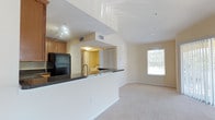 This single-story residence offers 2 bedrooms and 2 bathrooms within an open-concept floor plan designed for functional everyday living. The kitchen is equipped with warm wood cabinetry, dark granite countertops, a black refrigerator, and a built-in sink along a peninsula that serves as a natural divider between the kitchen and the living area. Track lighting illuminates the kitchen workspace, while recessed lighting throughout maintains a bright, consistent ambiance.

The living area features plush carpeting that adds a comfortable, understated feel to the space. A window along the far wall allows natural light to filter in, offering a view of the surrounding exterior. The open sightlines between the kitchen and living room create a sense of spaciousness and ease of movement throughout the main living area.

Visible from the main living space is a hallway leading to a bedroom with louvered closet doors, hinting at practical storage solutions. Neutral wall tones throughout contribute to a clean, versatile interior that can accommodate a range of furnishing styles. The layout balances connectivity between spaces while maintaining defined functional zones.