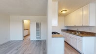 This single-floor residence offers a clean, modern aesthetic highlighted by light wood-style flooring that flows seamlessly throughout the entire space. The kitchen serves as a functional centerpiece, featuring crisp white shaker-style cabinetry, granite-look countertops, a gas cooktop, a stainless steel sink, and ample upper and lower storage. The layout includes a peninsula-style counter that helps define the kitchen area while maintaining an open connection to the adjoining living space. The living and bedroom areas are generously proportioned, bathed in natural light from large windows dressed with vertical blinds, and finished with the same consistent light flooring that gives the space a cohesive, airy feel. Vaulted ceiling angles in portions of the space add architectural interest and a sense of openness. The bathroom is well-appointed with a granite-topped vanity, white cabinetry, a large mirror, and a full tub-shower combo with a bright window above for natural light. A built-in wall vent provides climate control throughout. The overall palette of whites and neutrals creates a bright, clean environment that is both practical and inviting, making this a comfortable and move-in ready single-level home.